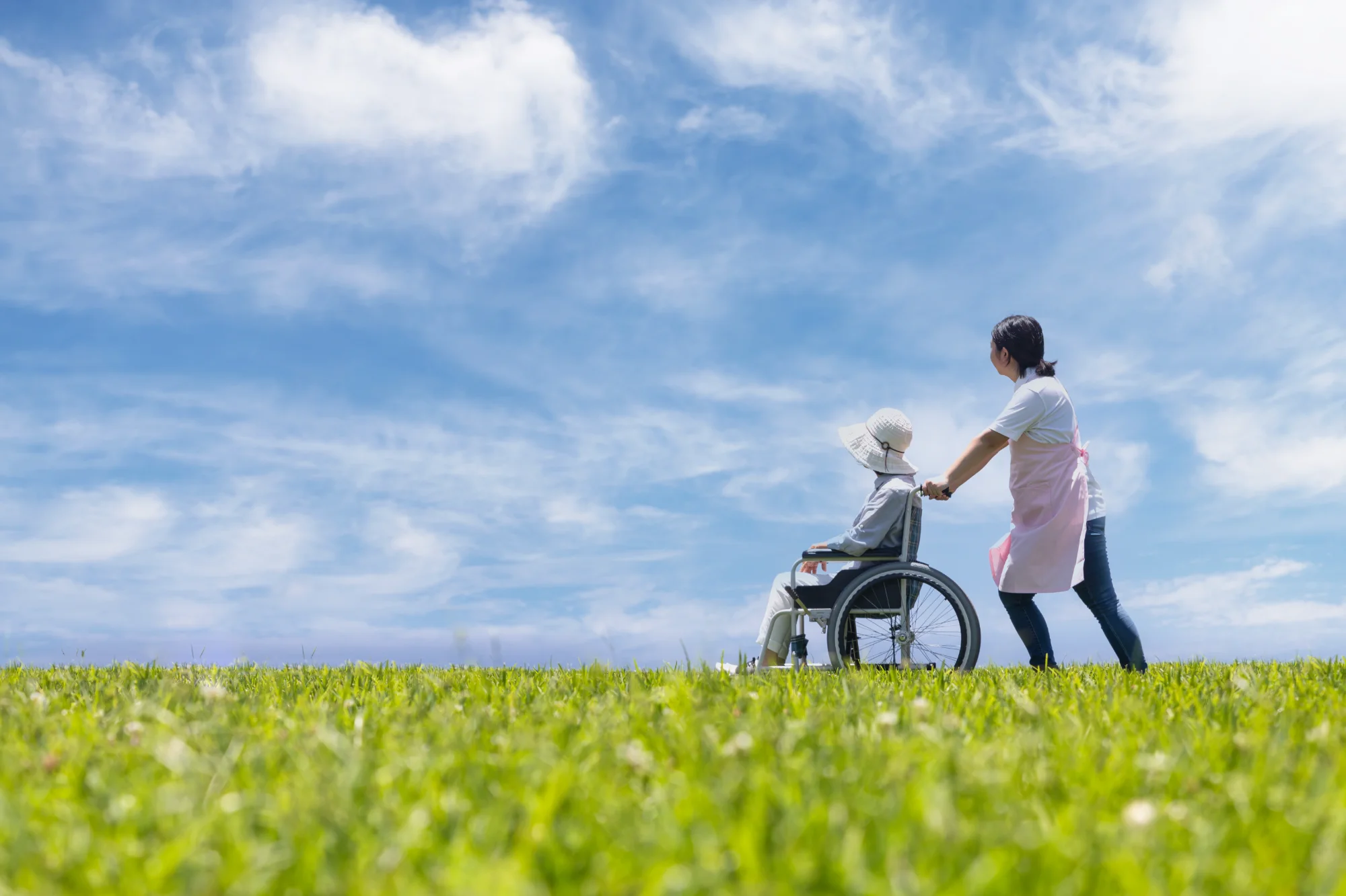 青空の下、介護士が車椅子の利用者様と散歩している様子